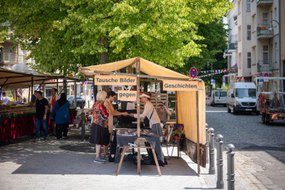 Holzpavillons mit beige-farbigem Sonnensegel auf einem Platz, wo Künstler ihre Werke ausstellen und mit Besuchern ins Gespräch kommen