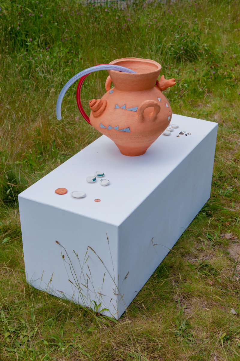 text: >
  Terrakottakrug mit blauem und rotem
  Henkel auf weißem Sockel, umgeben von
  verstreuten Münzen, Installation
  „Depot / 52.425678, 13.460060“ von
  Elisa Dierson und Katja Marie Voigt
decorative: false
source: manual