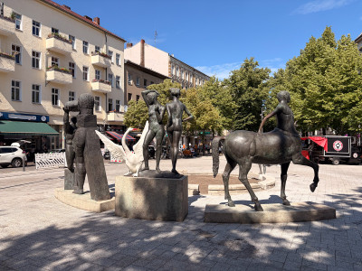Bronzeskulpturengruppe auf einem Platz zeigt mehrere menschliche und tierische Figuren in verschiedenen Posen, darunter zwei Frauen mit weißem Schwan und ein Zentaur