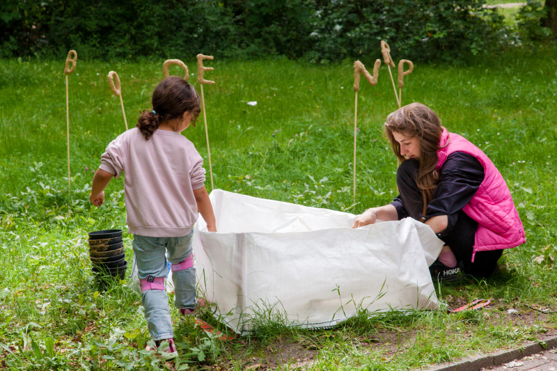 text: >
  Zwei Personen halten ein weißes Tuch
  über Gartenpflanzen, während Stöcke
  mit Buchstaben im Hintergrund den
  Workshop „Boden-Probe(n) – Playing
  in the Dirt!