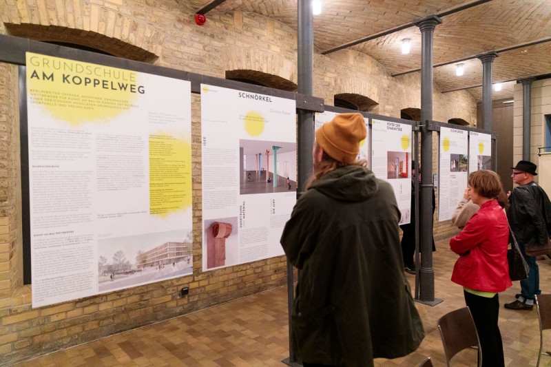 Besucherinnen und Besucher lesen ein Kunst am Bau Plakat in einer Ausstellung