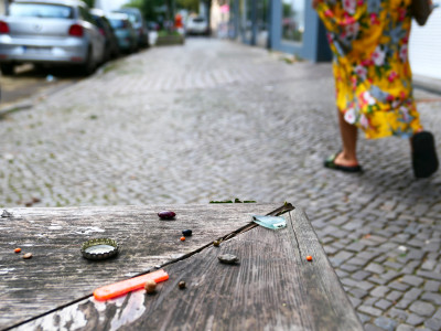 Oben rechts im Bild läuft eine Person, deren Beine nur zu sehen sind, auf dem Fußweg - links unten ist ein Holzbank mit kleinen Gegenständen drauf