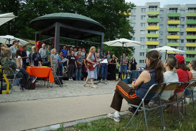 text: >
  Offene Chorprobe des Chors der Statistik
  im Freien auf dem Pavillonplatz vor
  einem Wohngebäude, dirigentin
  Bernadette La Hengst mit roter Gitarre
  steht unter einem Pavillon vor einer
  Gruppe von Zuschauern
decorative: false
source: reviewed