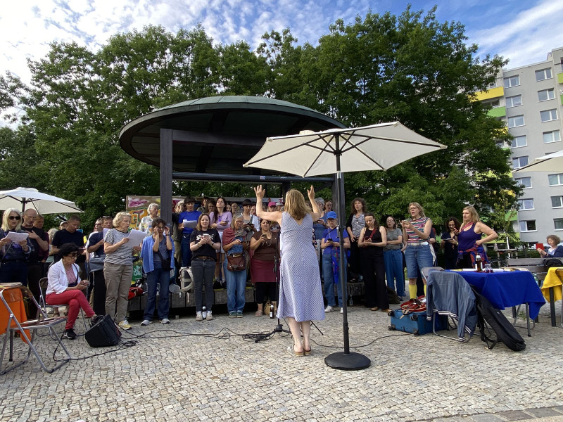 text: 'Dirigentin Bernadette La Hengst leitet eine Gruppe von Chorsänger*innen des Chors der Statistik unter einem Pavillon bei einer öffentlichen Chorprobe and der Pavillonplatz'
decorative: false
source: reviewed
